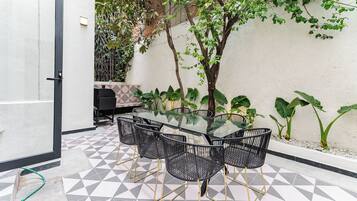A patio with a glass-top table surrounded by black chairs, a tiled floor, and a white wall with greenery.