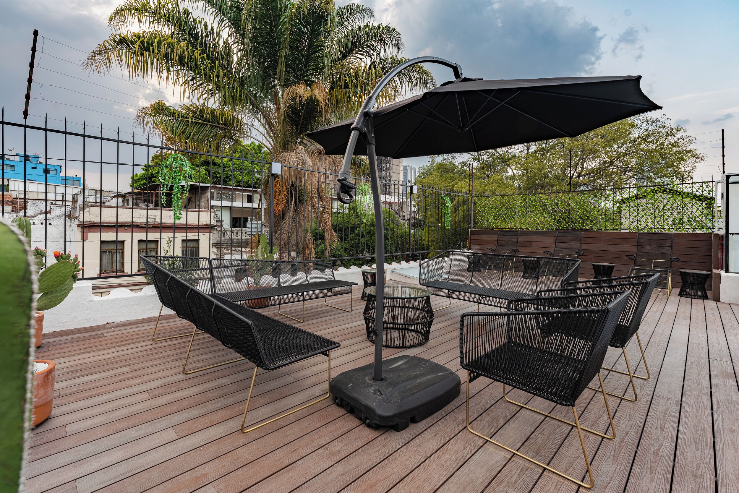 An outdoor patio with black metal chairs and a large umbrella, surrounded by a wooden deck and palm trees.