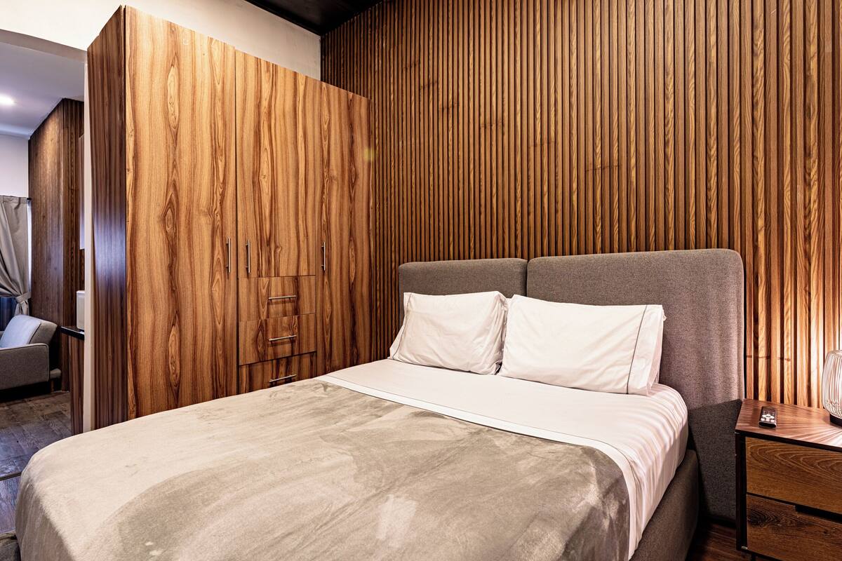 Een moderne slaapkamer met een wand van houten panelen, een bed met een grijze hoofdbord en een nachtkastje met een lamp.