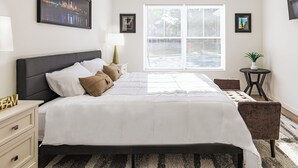Ein modernes Schlafzimmer mit einem großen Bett, einem Nachttisch, einer Nachttischlampe, einem kleinen runden Tisch mit einer Pflanze und einem Fenster mit Jalousien.