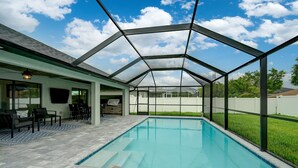 A covered outdoor swimming pool with a patio area, a covered seating area, and a view of a house in the background. - Modern Home with Fenced In Yard (Cape Coral)