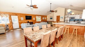 A spacious living area with a wooden floor, a dining table with chairs, a kitchen area with a bar, and a seating area with sofas. - Oceanside, Private Pool, Hot Tub, OS4N, Kaerolla Dawn (Corolla)