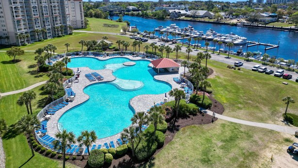 A large, irregularly shaped swimming pool surrounded by palm trees and lounge chairs, with a marina and boats in the background.