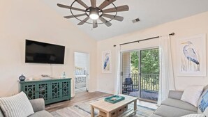 A spacious living room with a ceiling fan, a flat-screen TV, a green console table, and a sliding glass door leading to an outdoor area.