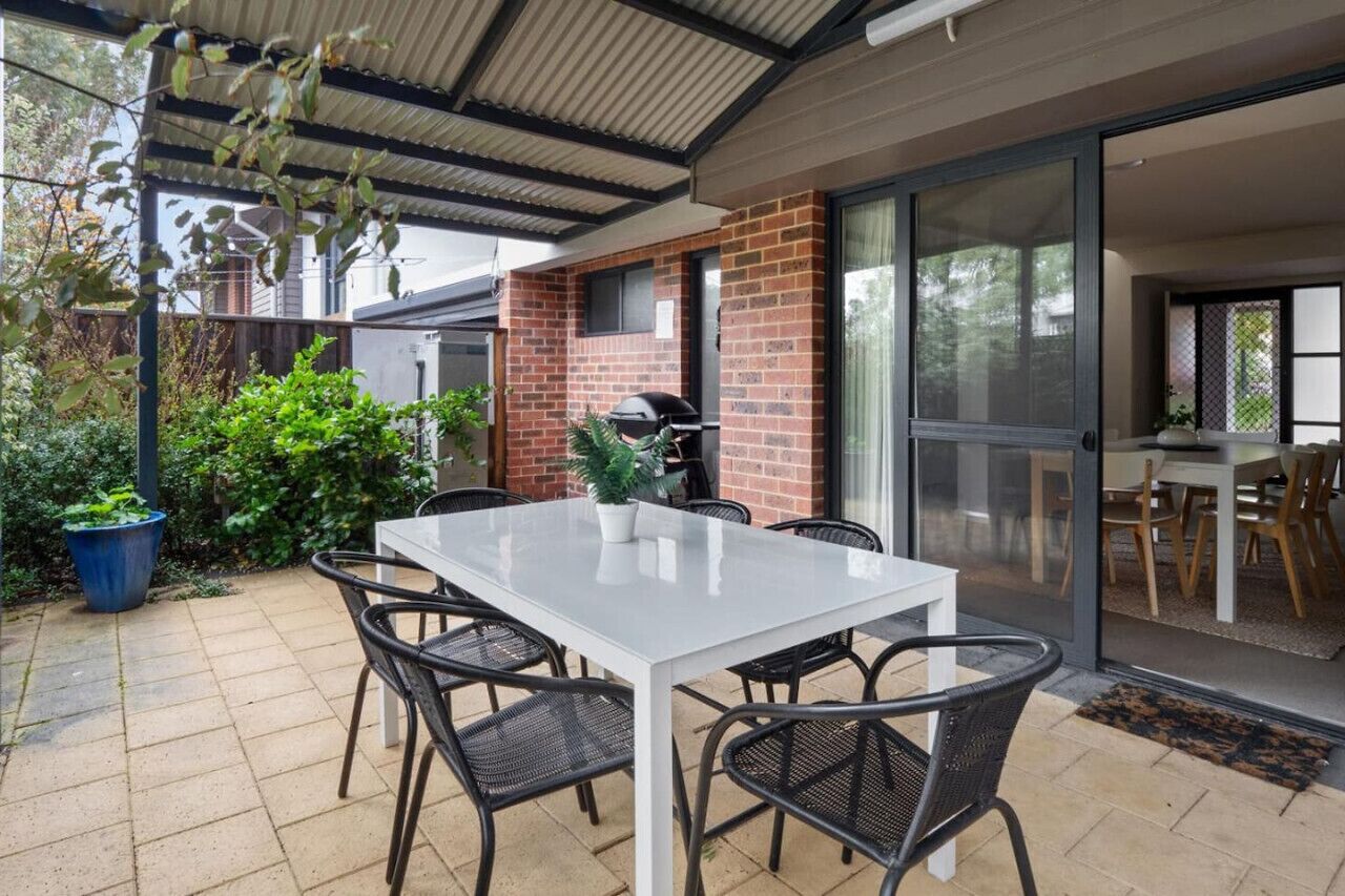 A covered patio area with a white table and black chairs, a brick wall, and a sliding glass door leading to a room.