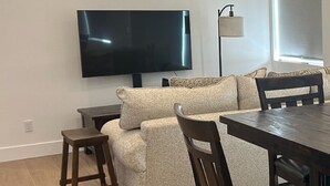 En moderne stue med et fladskærms-tv, en sofa, et spisebord og træstole.