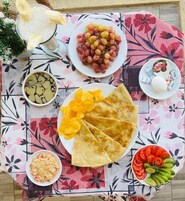 A table with a variety of food items including grapes, chips, a bowl of soup, a plate of sliced tomatoes and cucumbers, and a glass of a white beverage.