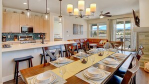A modern kitchen with a central island, dining area with a long table set for six, and a view of mountains through large windows.