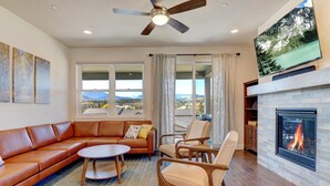 A living room with a leather sofa, a round coffee table, and two armchairs. A fireplace with a visible fire is on the wall, and there is a large window with a mountain view.