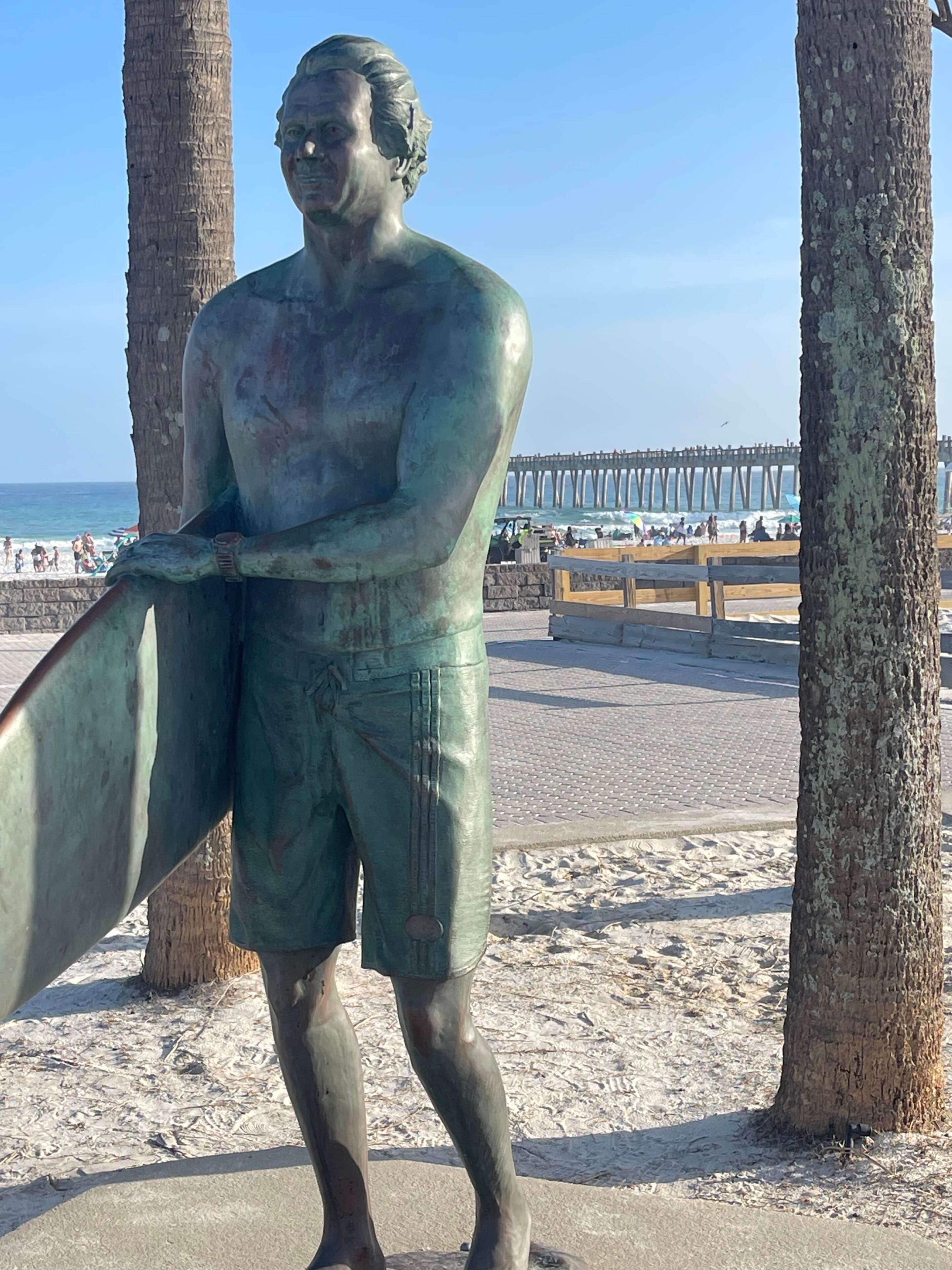 Uma estátua de bronze de um homem segurando uma prancha de surf, situada numa praia de areia com palmeiras e um cais ao fundo.