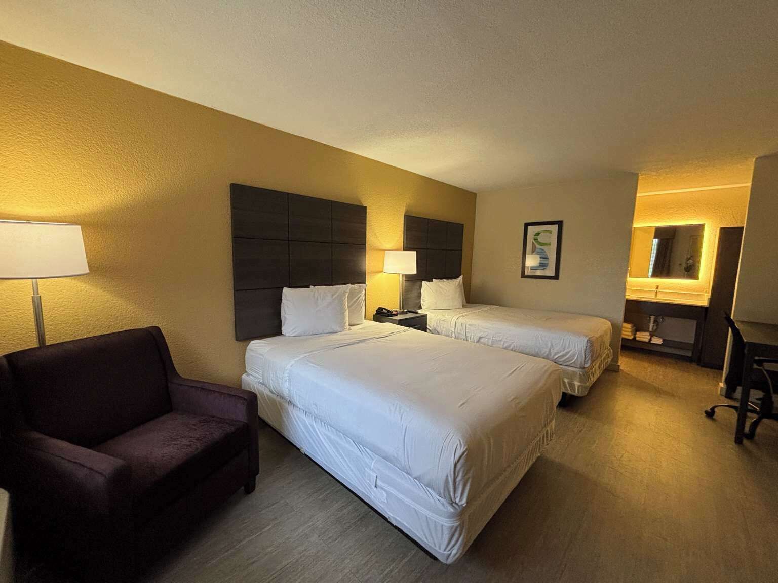 A hotel room with two beds, a desk, a chair, and a lamp.