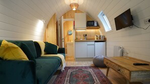 A cozy room with a green sofa, a wooden table, a kitchen area with a microwave, and a TV mounted on the wall. - Cosy Couples Cabin in the Scottish Highlands (Tomintoul)