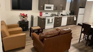 A modern apartment with a living room featuring a brown leather sofa and a beige armchair, a kitchen with dark wooden cabinets and white appliances, and a flat-screen TV mounted on the wall. - Bright Private 2BR Suite | Near Niagara Falls | Family-Friendly (Pelham)