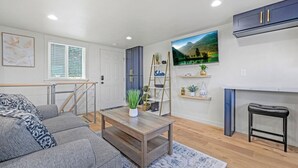 A modern living room with a grey sofa, a wooden coffee table, and a black stool. There is a staircase with a metal railing leading to a door. A TV is mounted on the wall above a shelf with decorative items. - Rucker Avenue Cozy Bungalow (Everett)
