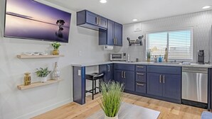 A modern kitchen with dark blue cabinets, a stainless steel dishwasher, and a large window with blinds. - Rucker Avenue Cozy Bungalow (Everett)