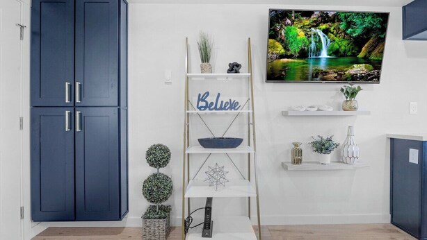 A modern living room with a TV mounted on the wall, a blue wardrobe, a white shelving unit with decorative items, and a small plant. - Rucker Avenue Cozy Bungalow (Everett)