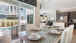 A modern dining area with a glass table, white chairs, and a view of a pool and blue buildings.