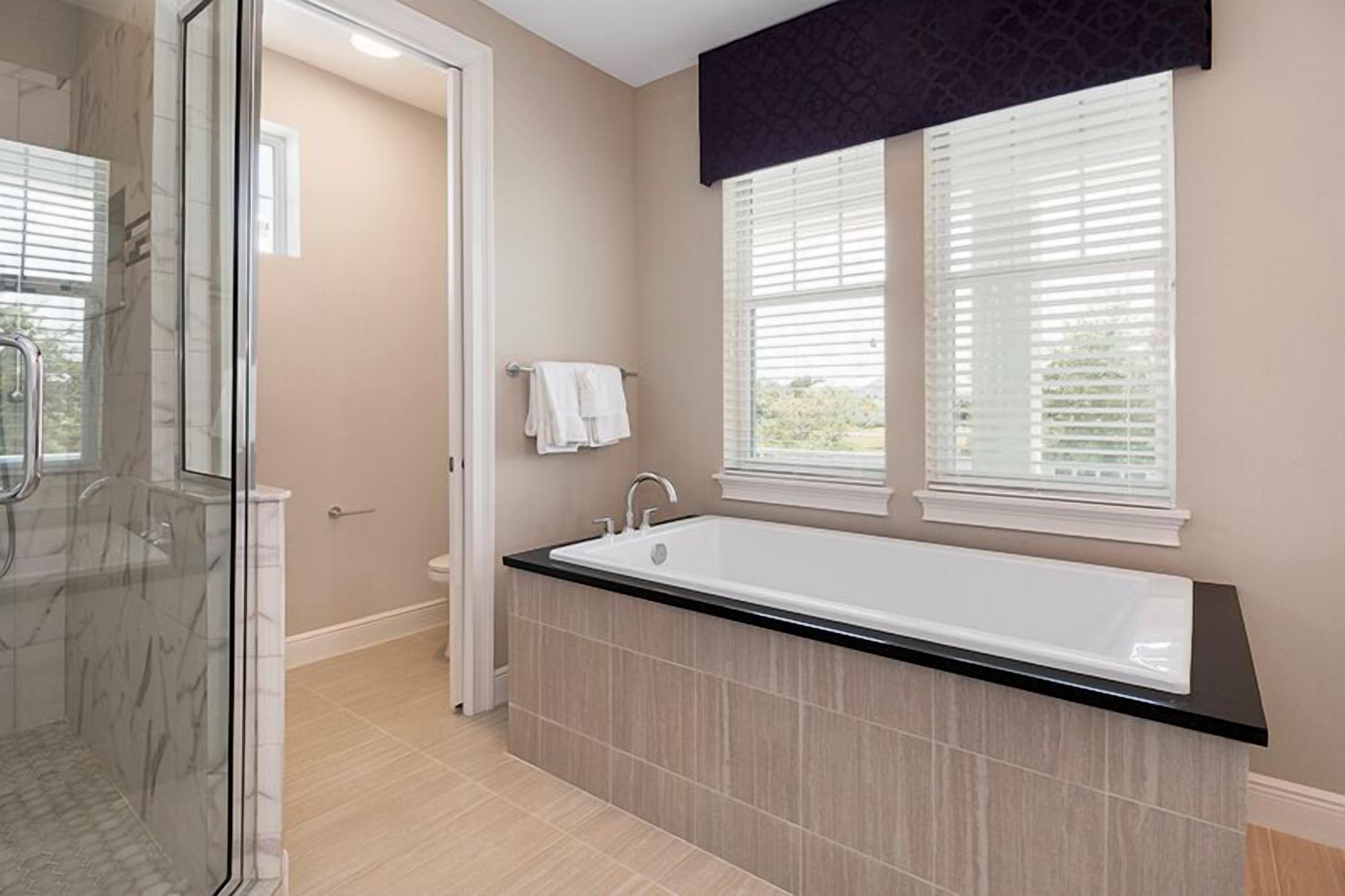 Un baño moderno con cabina de ducha de vidrio, una bañera grande y dos ventanas con persianas.