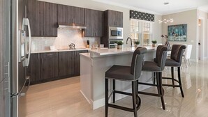 A modern kitchen with dark wood cabinets, a large island with bar stools, a stainless steel refrigerator, and a microwave.