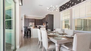 A modern dining area with a glass table, white chairs, and a kitchen in the background.
