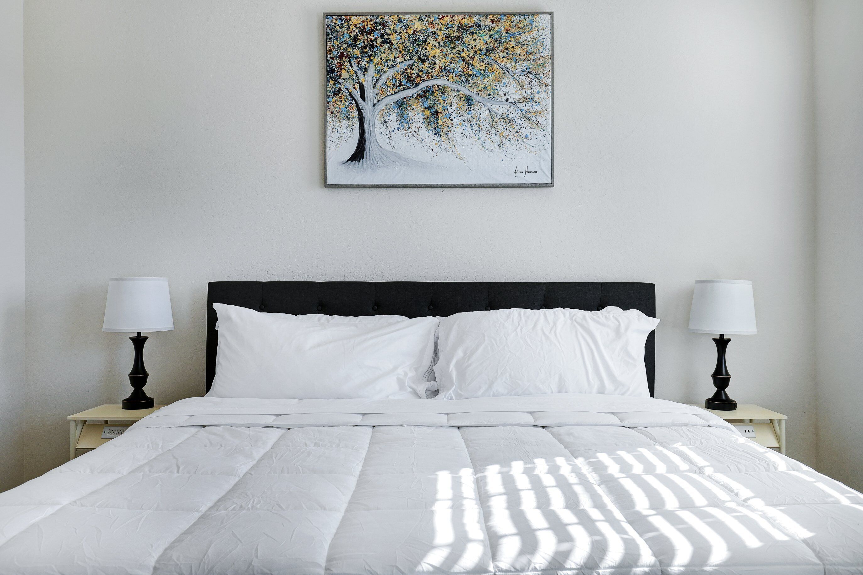 Una cama bien hecha con sábanas blancas, dos mesitas de noche con lámparas, y un cuadro con una imagen de un árbol en la pared.