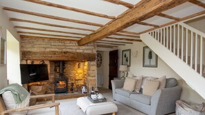 Ein gemütliches Wohnzimmer mit Kamin aus Stein, Holzbalken, einem grauen Sofa und einer Treppe.