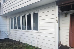 A white house with a number '13' on the door, a window with blinds, and a small garden area with a hose and a metal fence.