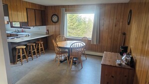 Een keuken met houten kastjes, een eettafel met stoelen, een klok en een raam met uitzicht op bomen.