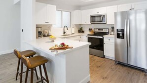 A modern kitchen with white cabinetry, a stainless steel refrigerator, a microwave, and a built-in oven. There is a bar area with stools and a fruit bowl.