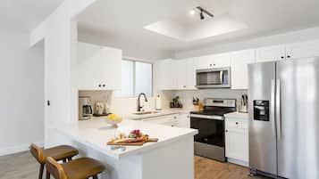 A modern kitchen with white cabinetry, a stainless steel refrigerator, a microwave, and a built-in oven. There is a bar area with stools and a fruit bowl.