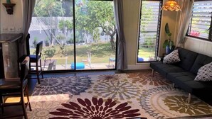 Un salon avec une grande fenêtre, un ventilateur de plafond, un canapé et un tapis à motifs.