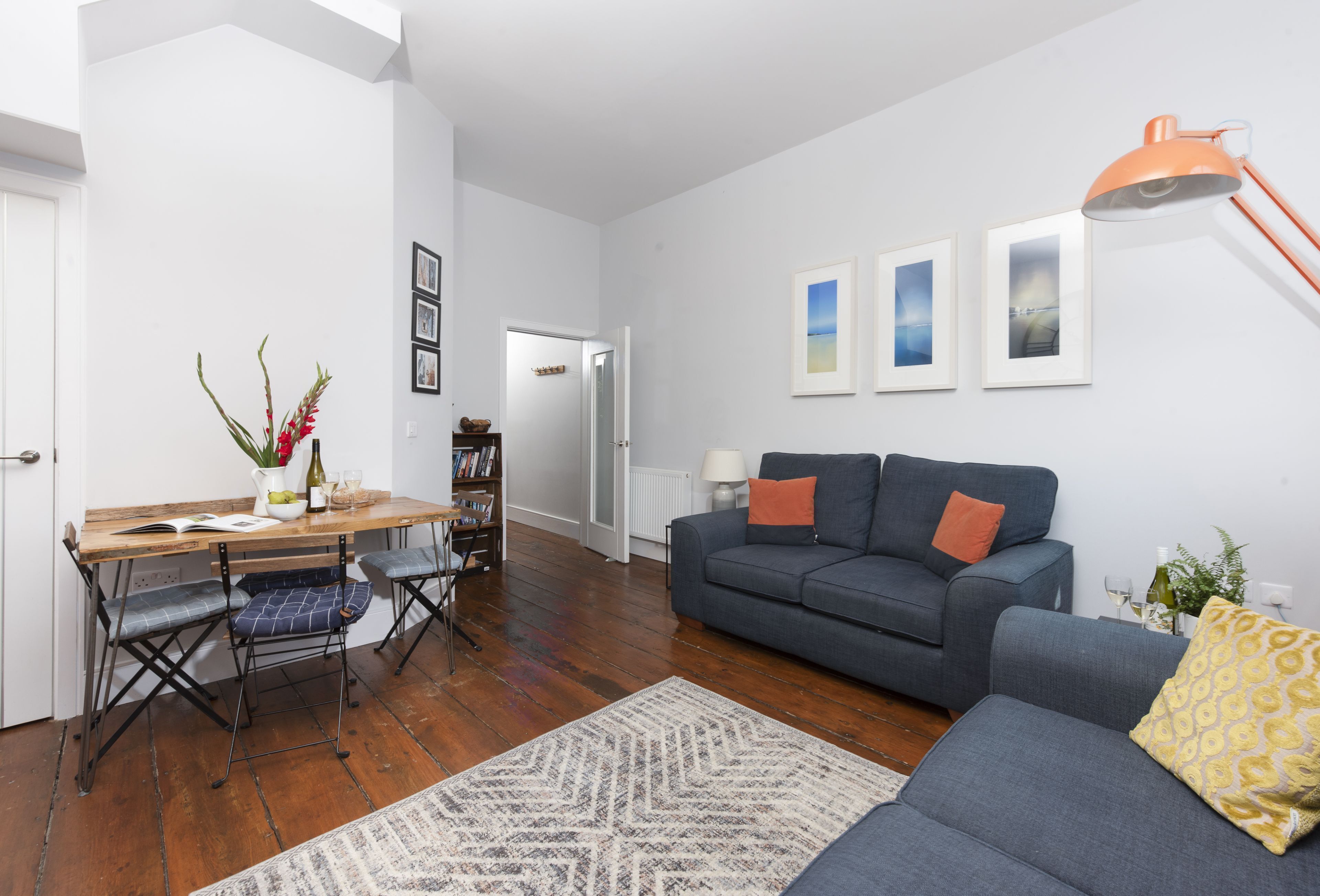 A living room with a grey sofa, a wooden dining table with blue cushioned chairs, a patterned rug, and framed pictures on the wall.