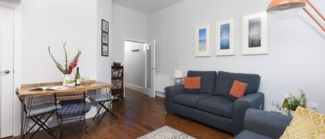 A living room with a grey sofa, a wooden dining table with blue cushioned chairs, a patterned rug, and framed pictures on the wall.