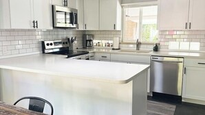 A modern kitchen with white cabinetry, a central island, a stainless steel dishwasher, and a dining table with a rustic wooden top. - Crown Hill Retreat - Hot Tub/Yard/Parking (Lakewood)