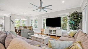 A spacious living room with a large sofa, a coffee table, and a television. There is a dining area with chairs and a table, and a ceiling fan.