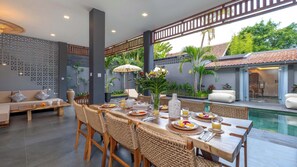 Un coin repas avec vue sur la piscine, comprenant une longue table en bois dressée pour le petit-déjeuner, des chaises en osier et un vase décoratif.