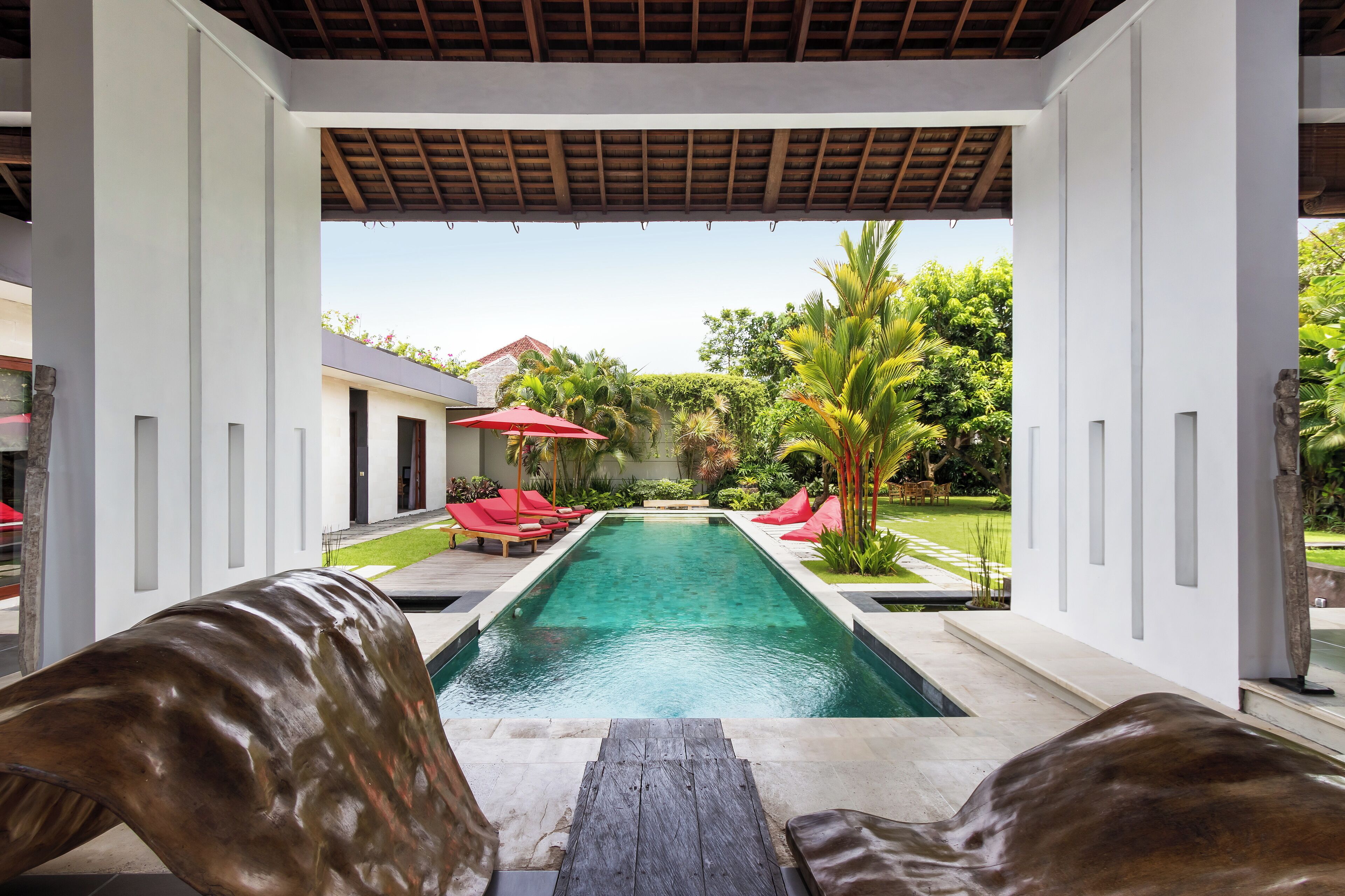 A pool area with a wooden deck, red lounge chairs, and a view of a house and garden.