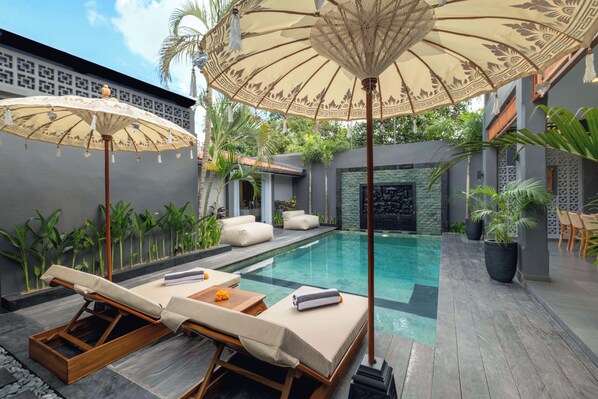 A modern outdoor pool area with lounge chairs and umbrellas, surrounded by plants and a stone wall. - Alarte Estate by Villa Finder (Seminyak)