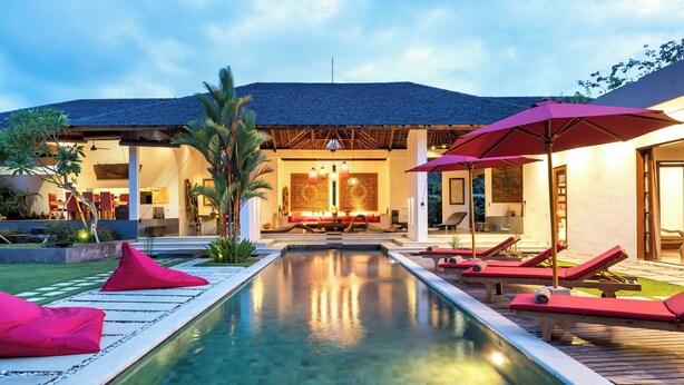 Un espace piscine avec des chaises longues et des parasols, un bâtiment bien éclairé avec un toit en tuiles et un ciel dégagé.