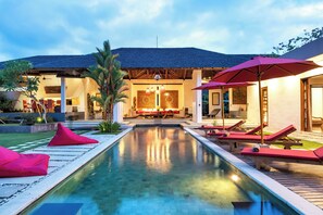 A pool area with lounge chairs and umbrellas, a well-lit building with a tiled roof, and a clear sky. - Alarte Estate by Villa Finder (Seminyak)