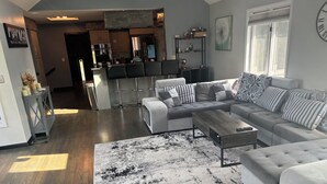 Ein modernes Wohnzimmer mit einem Ecksofa, einem Couchtisch und einem schwarz-weißen Teppich. Im Hintergrund ist eine Küche mit einer Bar und Barhocker zu sehen.