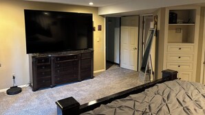 Ein Schlafzimmer mit einem großen Flachbildfernseher, einer dunklen Holzkommode, einem Bett mit grauer Steudecke, einem Deckenventilator und einem begehbaren Kleiderschrank.