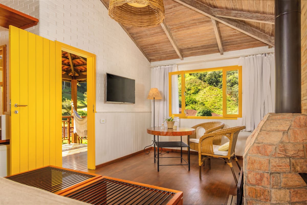 A room with a yellow door, a stone fireplace, a wicker chair, a table with a lamp, and a window with a view of greenery.