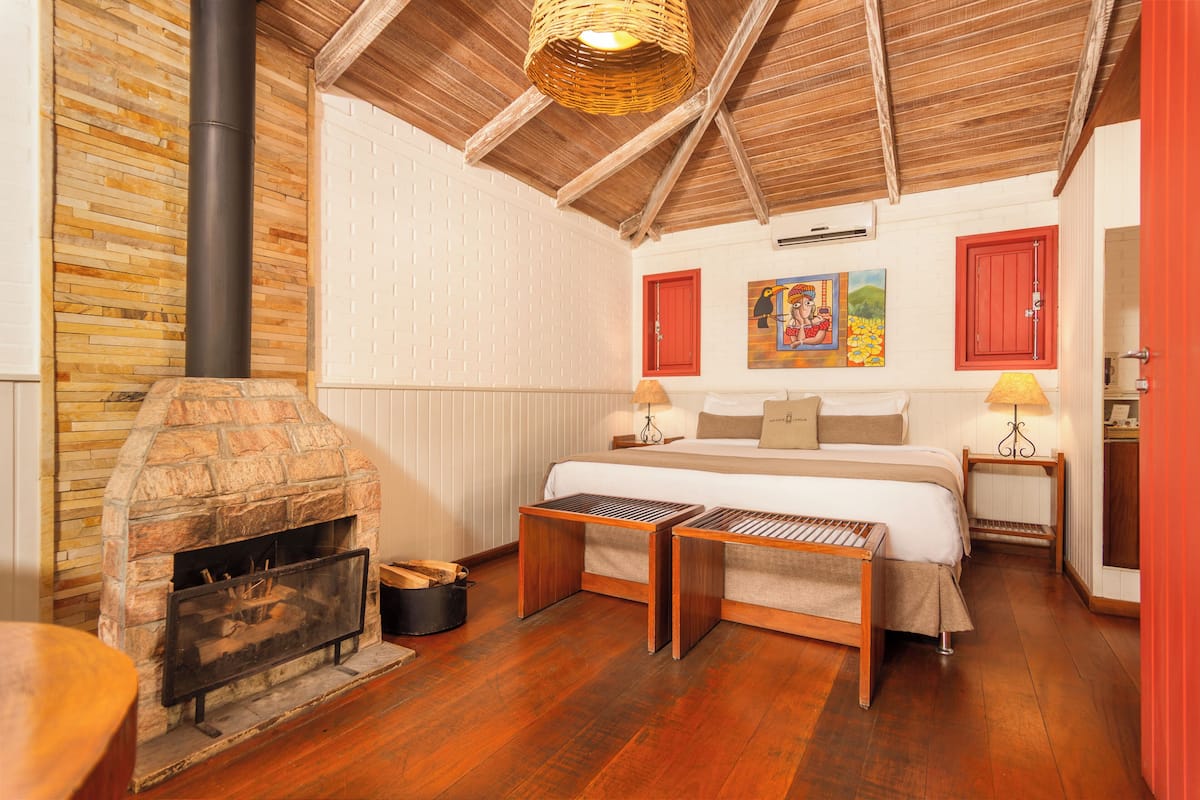A bedroom with a stone fireplace, wooden flooring, a bed with pillows, a nightstand with a lamp, and a painting on the wall.