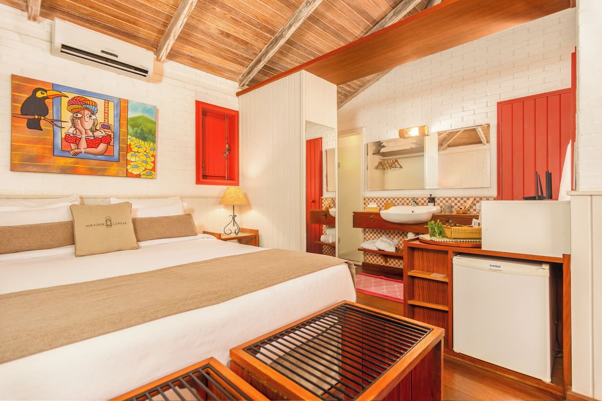 A hotel room with a bed, a bedside table with a lamp, a small wooden bench, a sink, and a refrigerator.