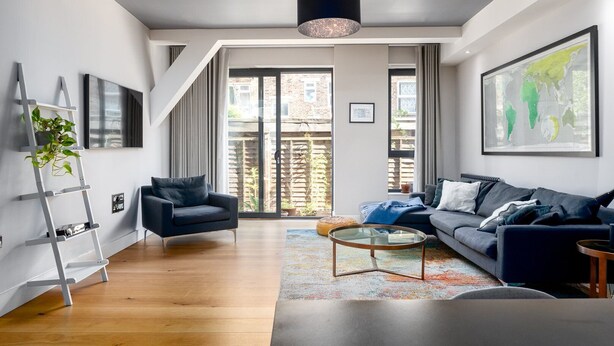 Una sala de estar moderna con un sofá, un sillón y una mesa de centro. Hay una escalera apoyada en la pared y un ventanal con vistas al exterior.