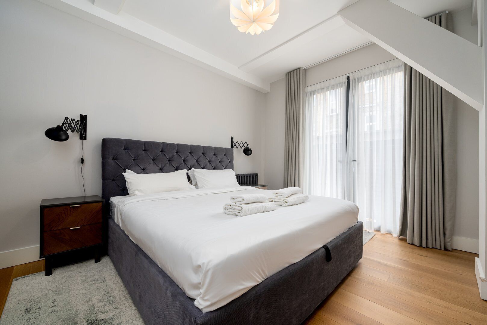 Een moderne slaapkamer met een groot bed, een nachtkastje, een lamp en uitzicht op het groen door dunne gordijnen.