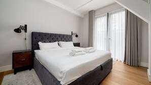 Un dormitorio moderno con una cama grande, una mesita de noche, una lámpara y vistas al exterior a través de cortinas translúcidas.