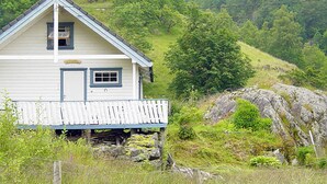 A white wooden house with a porch, surrounded by lush greenery and rocky terrain. - 5 Person Holiday Home in Akra (Kvinnherad)