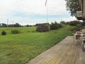 A grassy field with a flagpole, a paved area with chairs, and a building with a chimney.